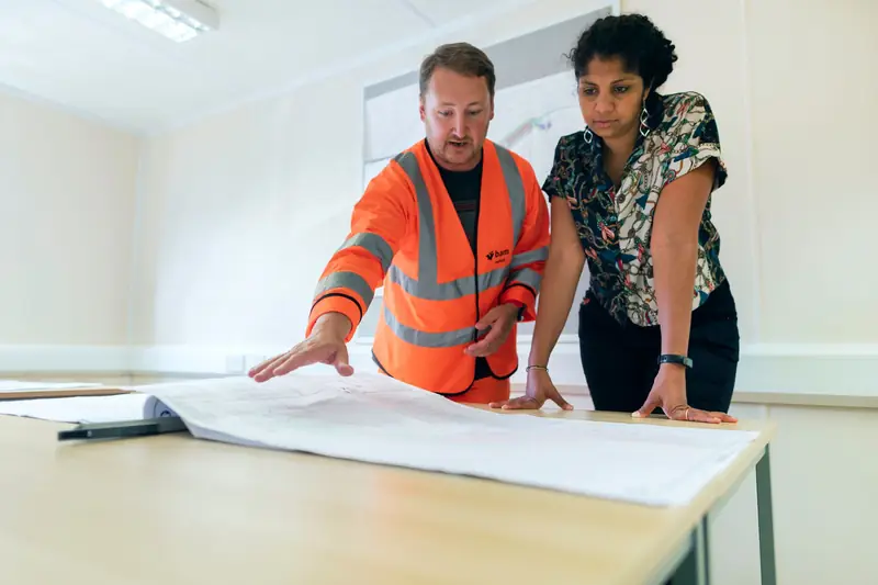 Two people in work clothes reviewing large printed plans at a table
